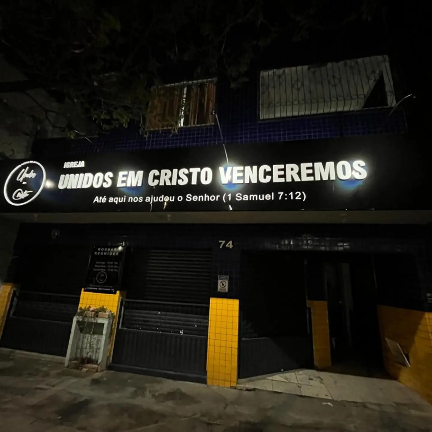 A mesma fachada à noite, com a letra caixa iluminada acesa e legível a quarteirões de distância