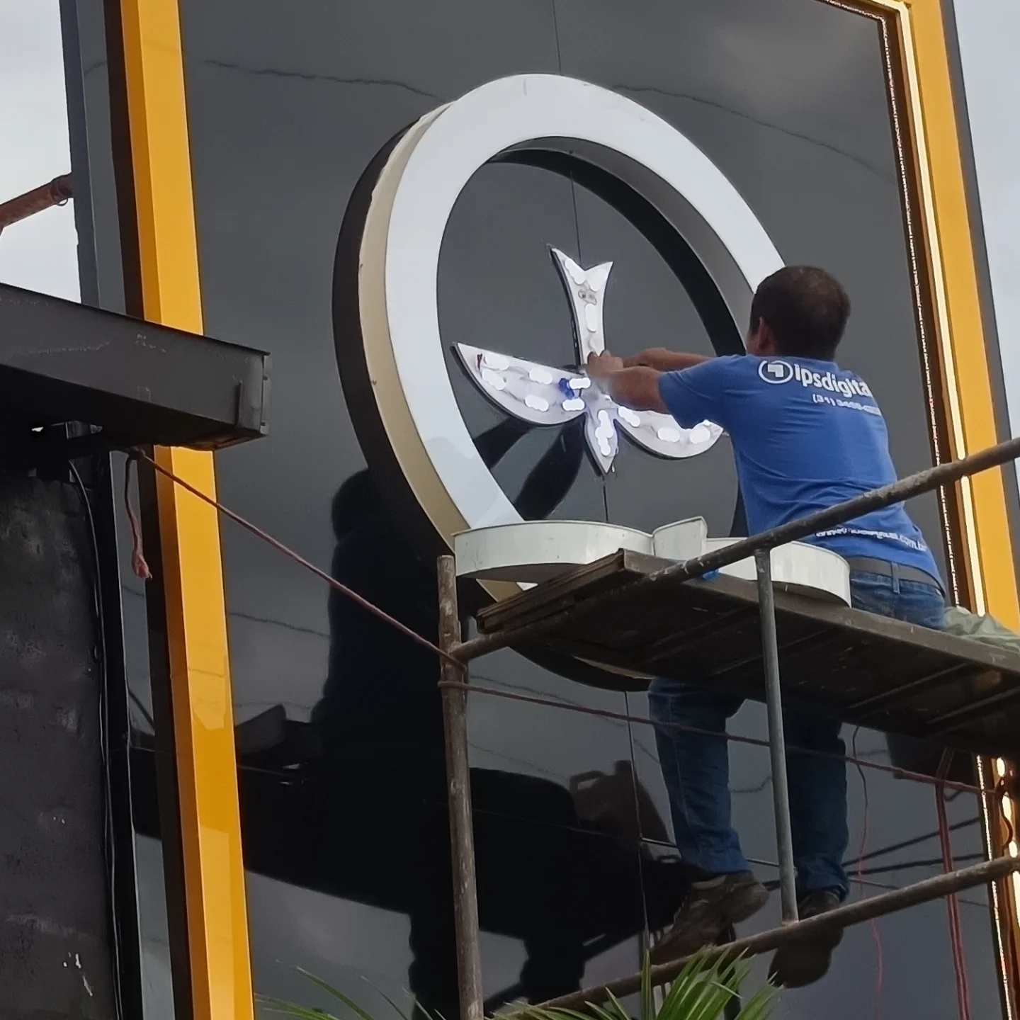 Técnico da IPS Digital, de camiseta da empresa, montando à mão o símbolo iluminado sobre o andaime