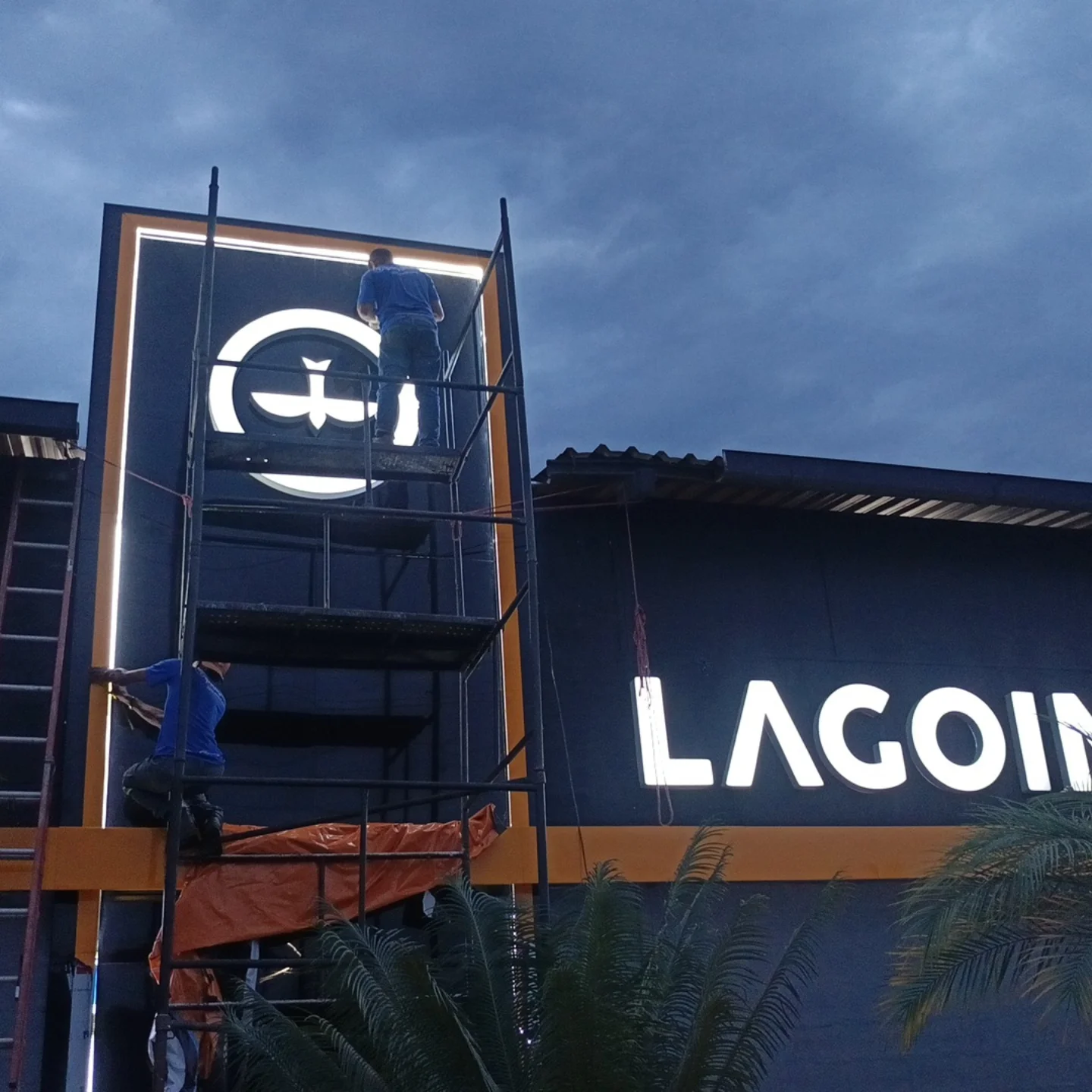 Dois técnicos no andaime ao entardecer, com as letras da fachada já acesas