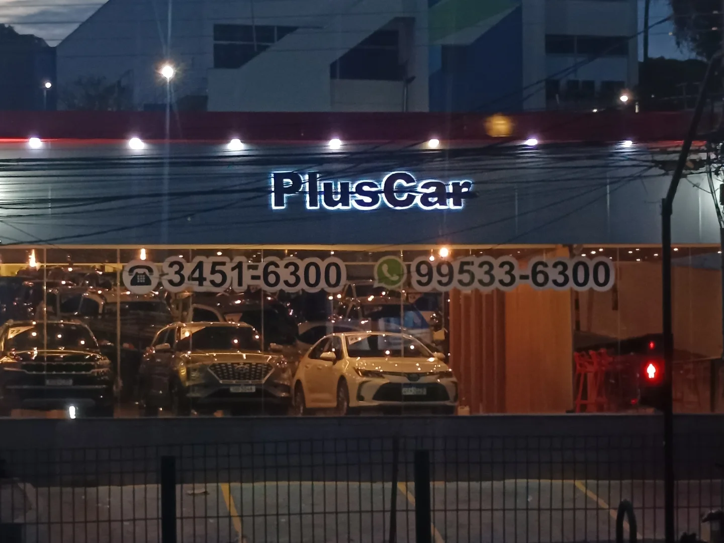 Fachada da revenda PlusCar com letra caixa iluminada e vitrine adesivada com telefone e WhatsApp